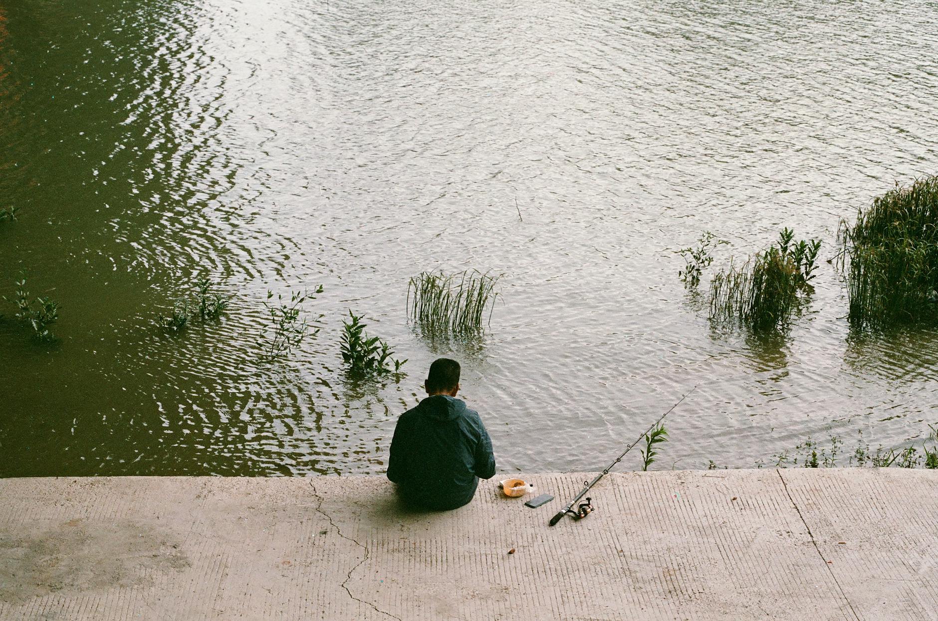 Samotny wędkarz siedzi nad rzeką w spokojnym, zielonym otoczeniu